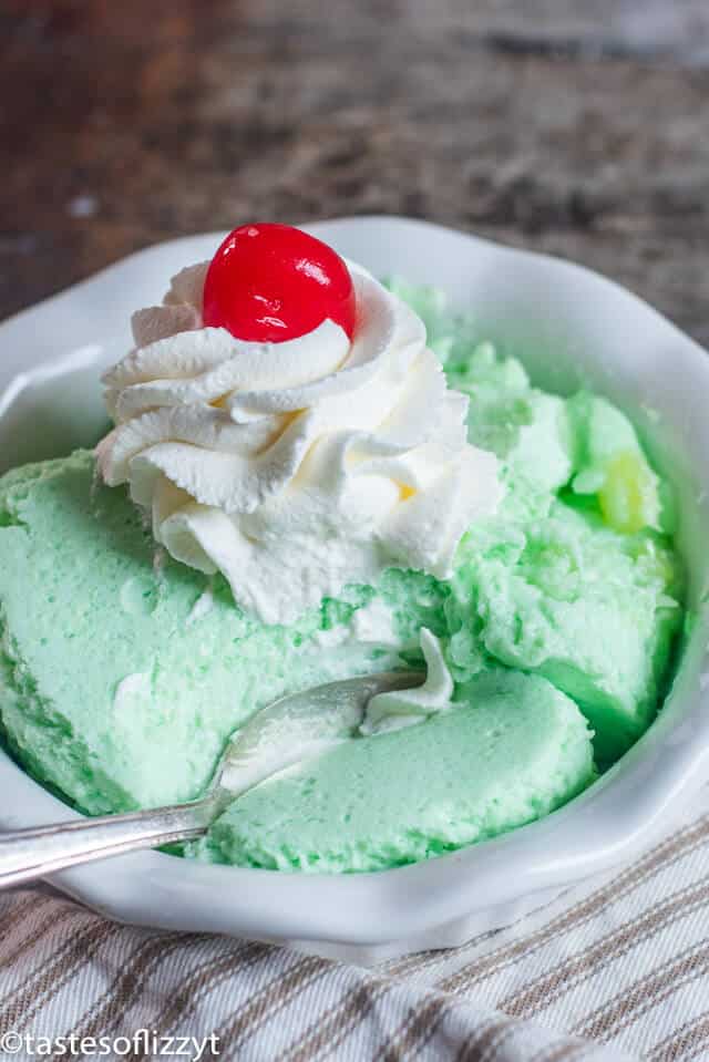 A bowl of lime jello salad with whipped cream and cherry