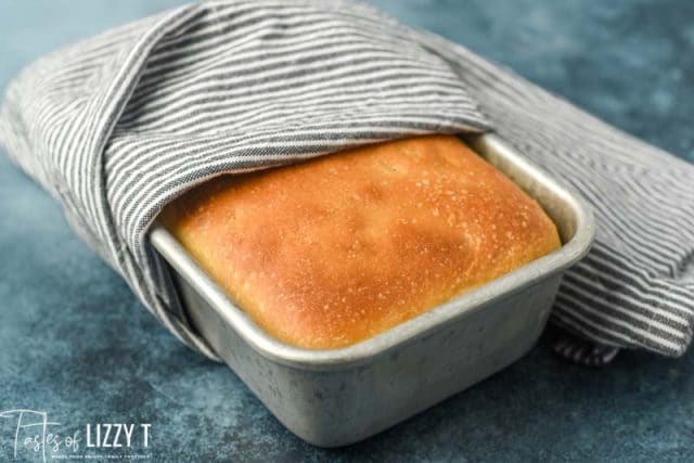 golden brown white bread in a loaf pan wrapped in a towel