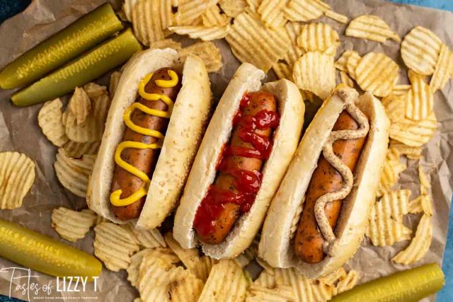 3 hot dogs in buns on brown paper with pickles and chips