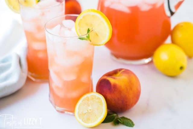 two cups of peach lemonade with ice and a pitcher in teh background