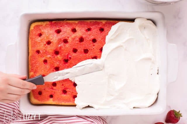 strawberry poke cake with spatula spreading white frosting on top