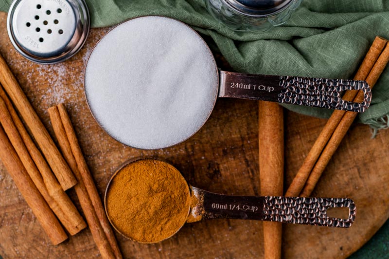 cinnamon and sugar in measuring cups on a table with cinnamon sticks