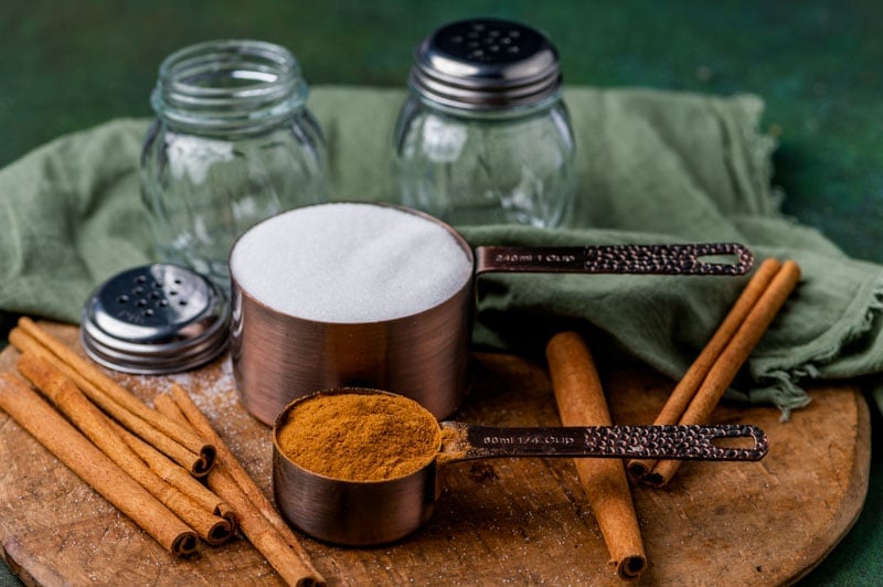 cinnamon and sugar in measuring cups on a table with cinnamon sticks