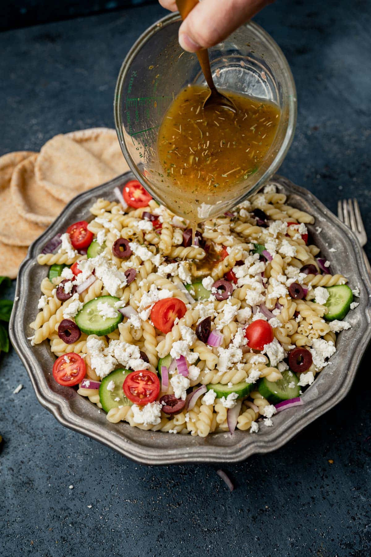 pouring greek salad dressing on pasta salad