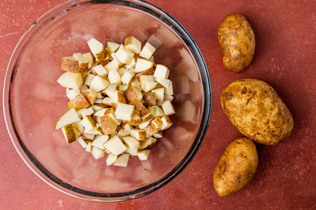 3 russet potatoes and a bowl of cubed potatoes on a table