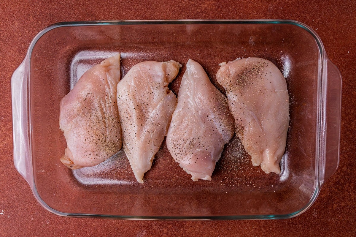 4 raw chicken breasts with salt and pepper sprinkled on top in a glass pan