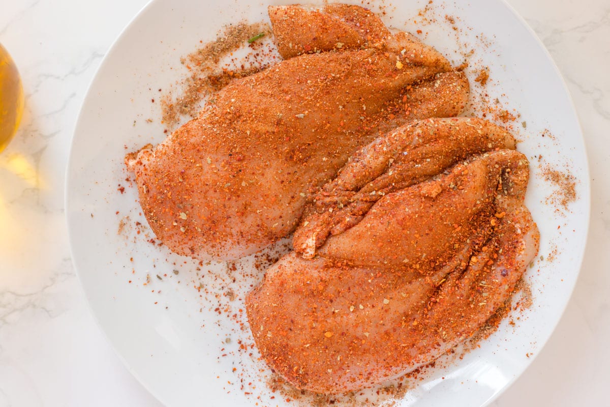 two uncooked chicken breasts on a plate with seasonings