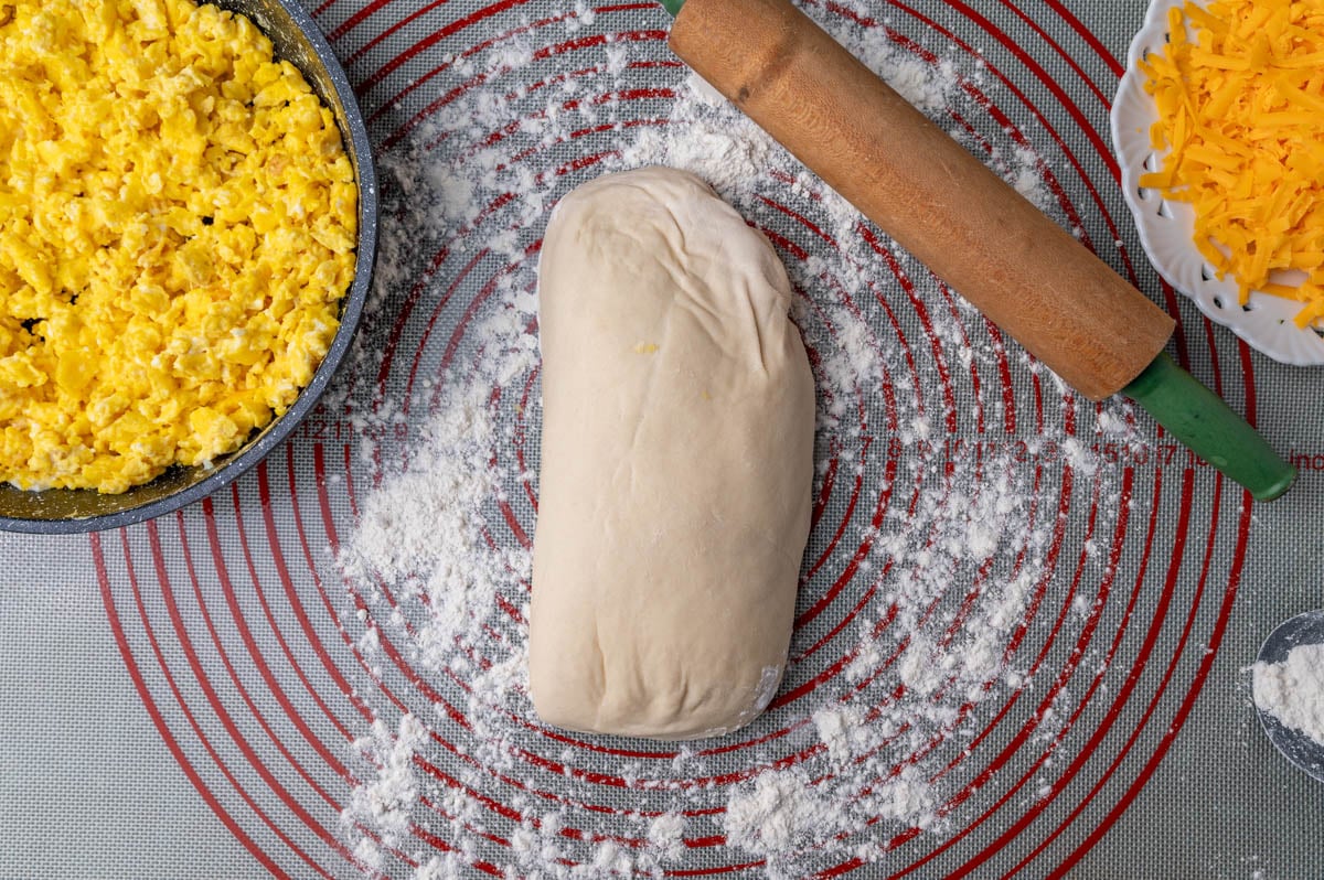 a loaf of bread dough on a pastry mat with rolling pin, eggs and cheese beside it