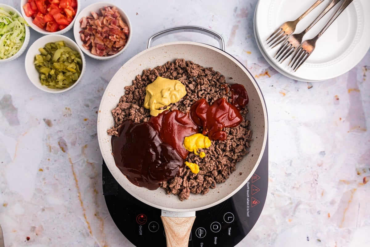 browned ground beef with ketchup, mustards and bbq sauce on top in a skillet