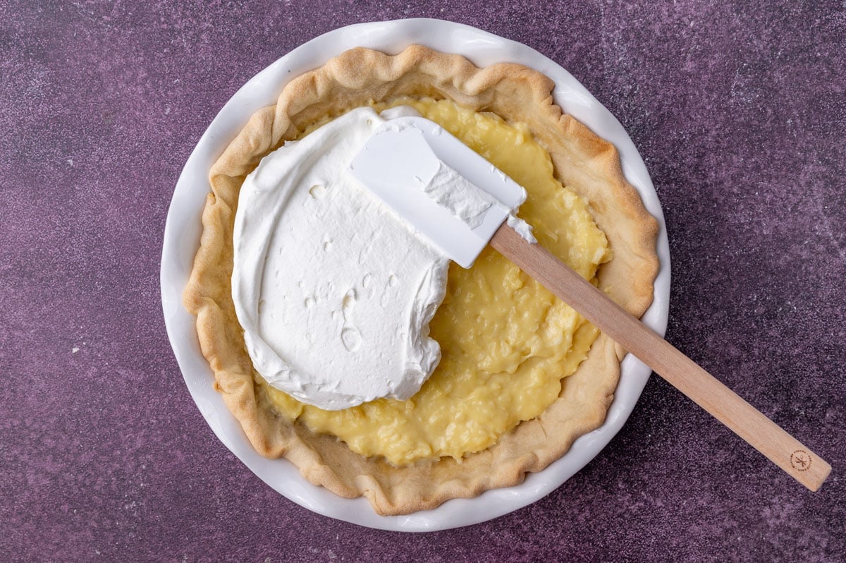 a spatula spreading whipped cream over coconut cream pie