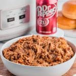 a bowl of pulled pork with dr pepper and a slow cooker in the background